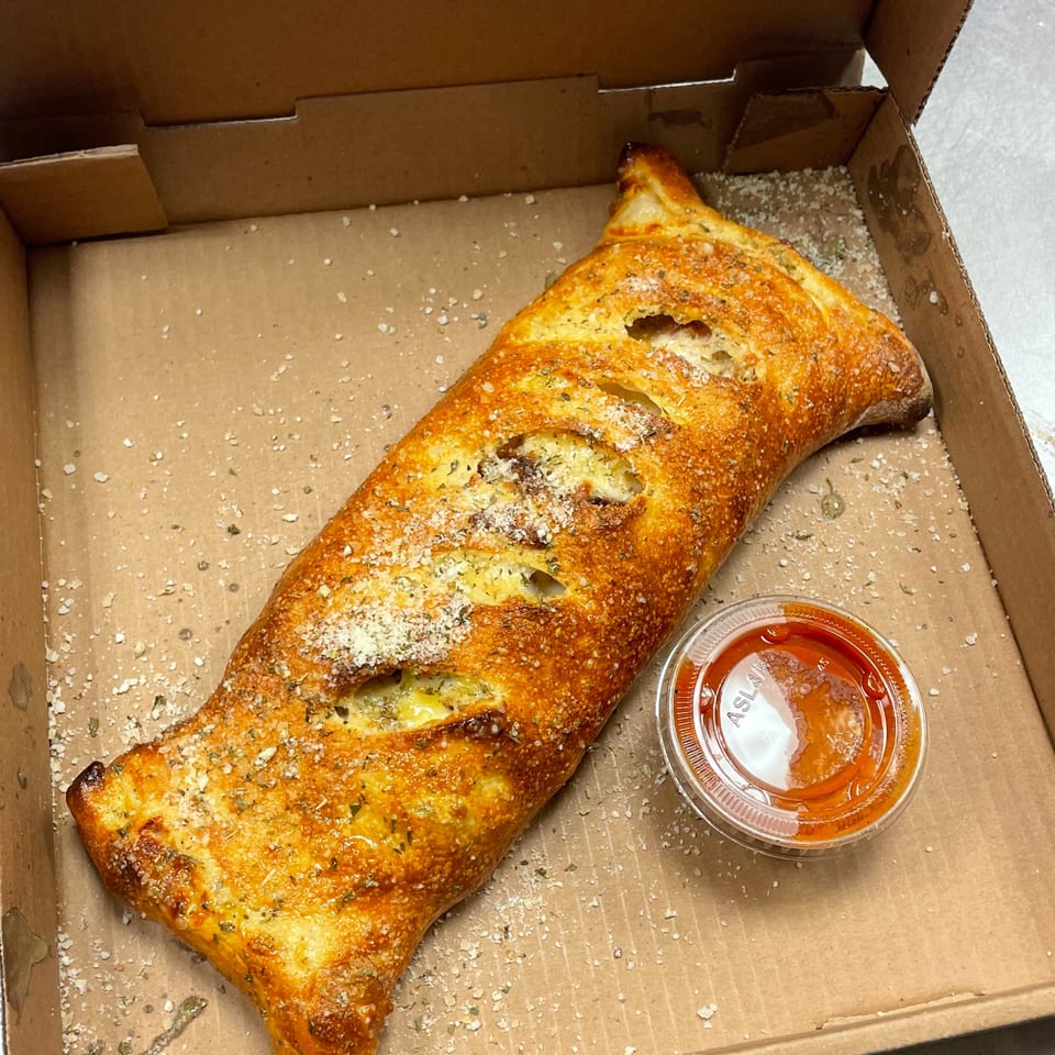 A golden-brown calzone, sprinkled with herbs and cheese, rests in a pizza box with a side of marinara.