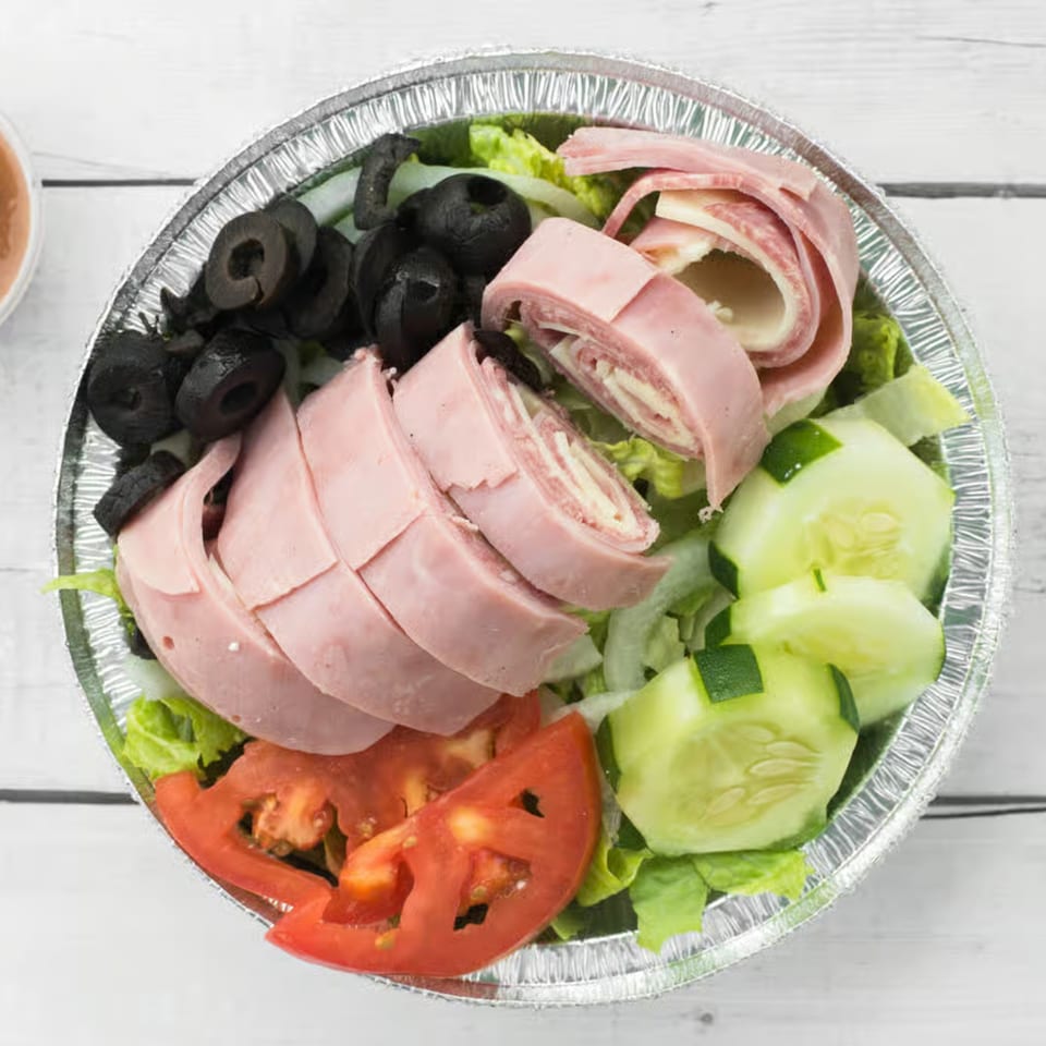 Antipasto salad with ham, salami, cheese rolls, black olives, tomatoes, and cucumbers in a foil bowl.