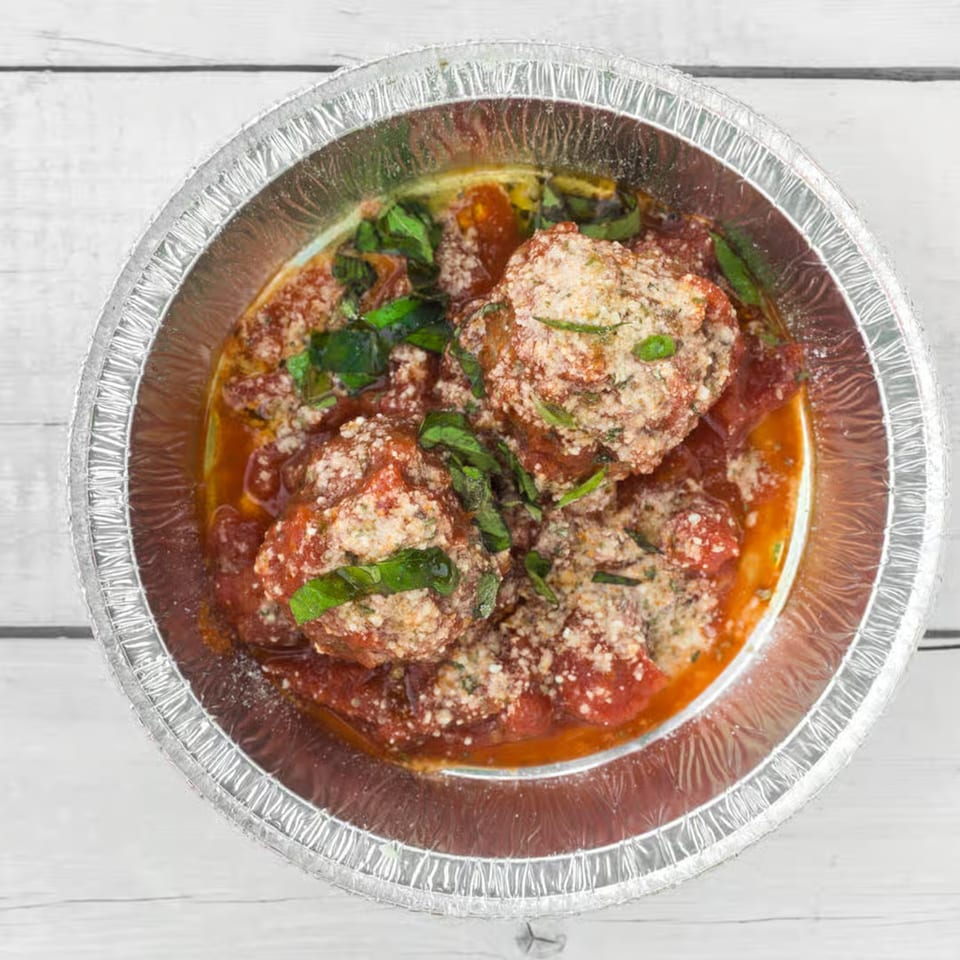 Meatballs in red sauce with parmesan cheese and basil in a foil container.